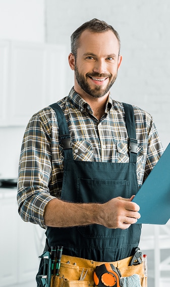 Homme souriant, ouvrier qualifié en salopette et chemise à carreaux, tenant un presse-papiers. Sa ceinture à outils est visible.