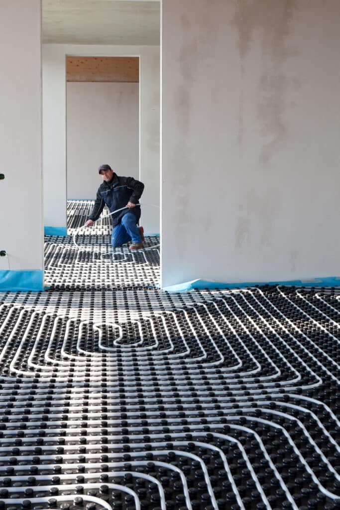 Ouvrier posant des tuyaux blancs de chauffage au sol sur un panneau isolant noir dans une pièce en chantier, murs clairs.