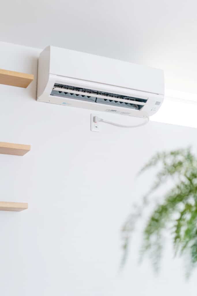 Climatiseur mural blanc branché sur une prise électrique 200V, près d'étagères en bois, dans un intérieur lumineux.