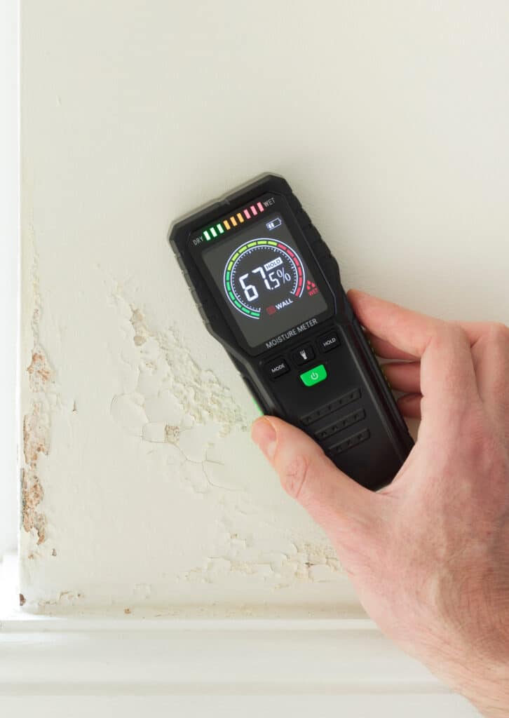 Main mesurant l'humidité d'un mur avec peinture écaillée. Le détecteur affiche 67.5% WET, indiquant un problème d'humidité.