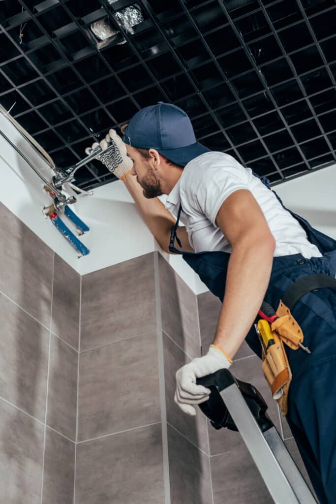 Plombier sur échelle, fixant des tuyaux au plafond suspendu à grille noire. Il porte un uniforme, des gants de travail et une casquette.