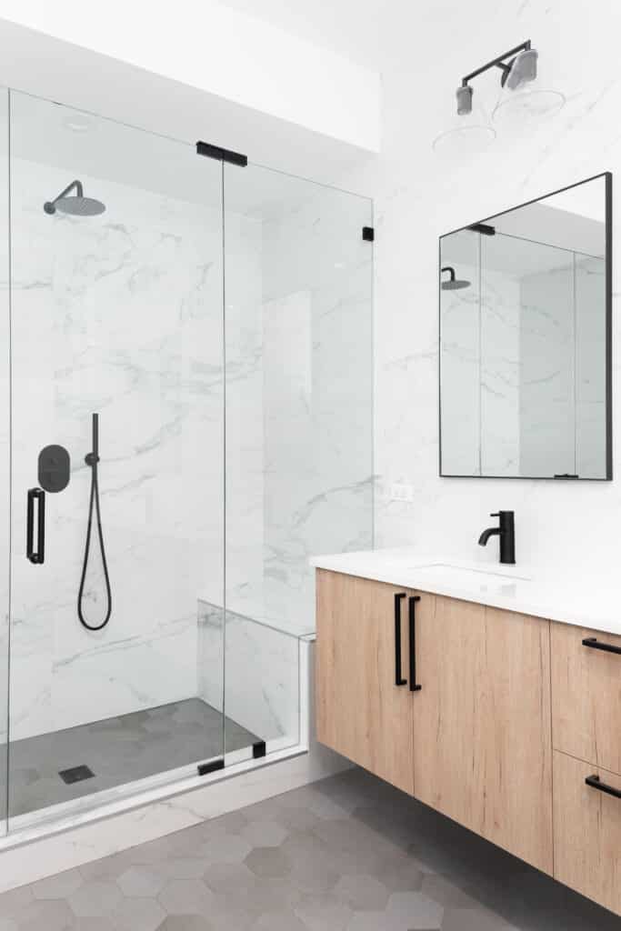 Salle de bain moderne avec douche à l'italienne en marbre blanc et verre, meuble vasque en bois clair, robinet noir mat et carrelage hexagonal gris.